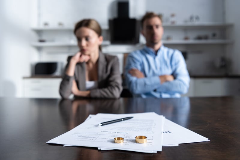 Blurred couple in background with divorce papers and wedding rings on kitchen counter in foreground