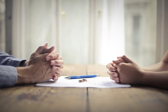 Two people sitting across from each other at a table with clasped hands, documents and a pen between them