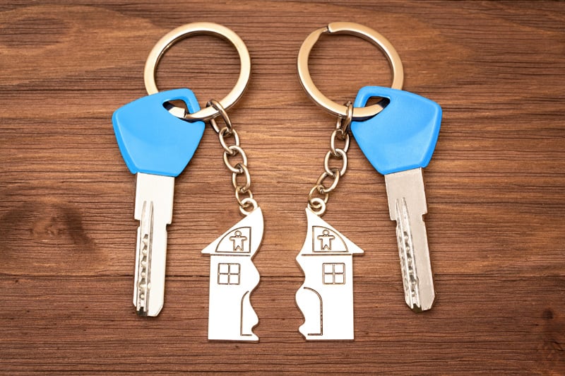 Two sets of keys with blue tops and broken house-shaped keychains split in half on wooden surface