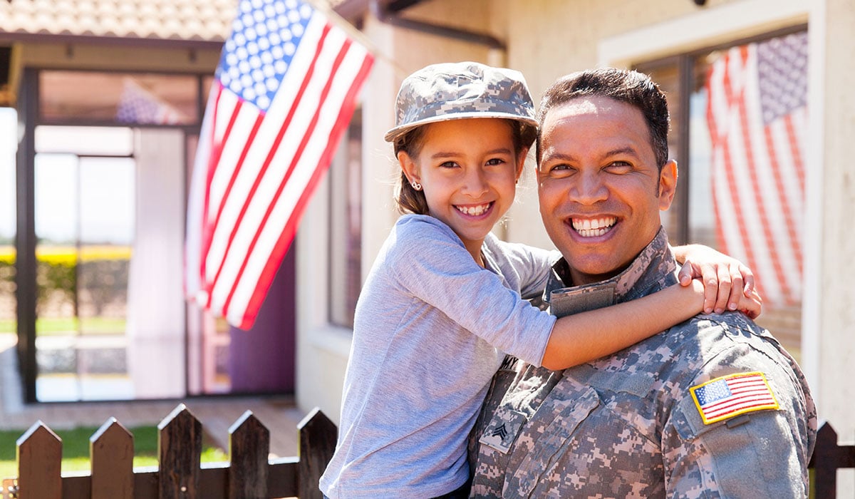 dad dressed in military uniforming holding smiling daughter in front of home