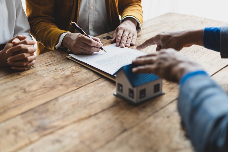 Hands signing documents at wooden table with small house model nearby during real estate transaction Hands signing documents at wooden table with small house model nearby during real estate transaction