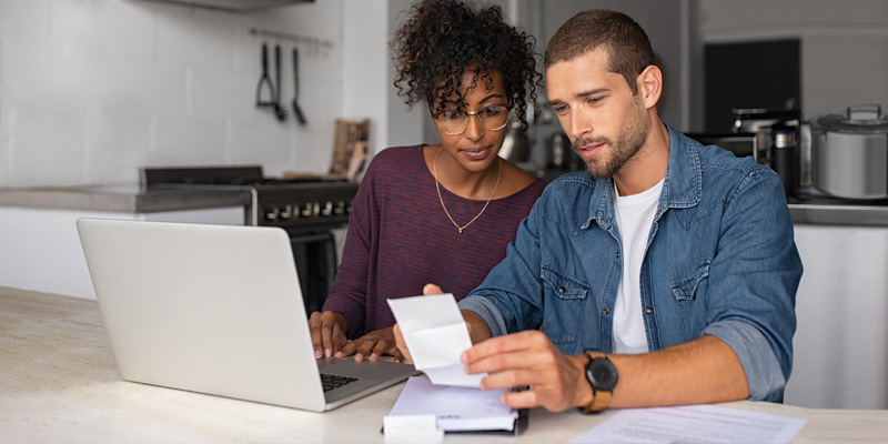 Young multiethnic couple examining home finance in their kitchen by reviewing paper bills and documents on their laptop